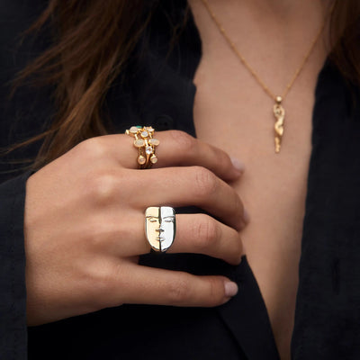 Close-up of a person wearing an Awe Inspired gold necklace and multiple rings. A prominent stack of Face to Face Rings is on their index finger, and a decorative nesting ring is on their pinky finger, over a black garment.