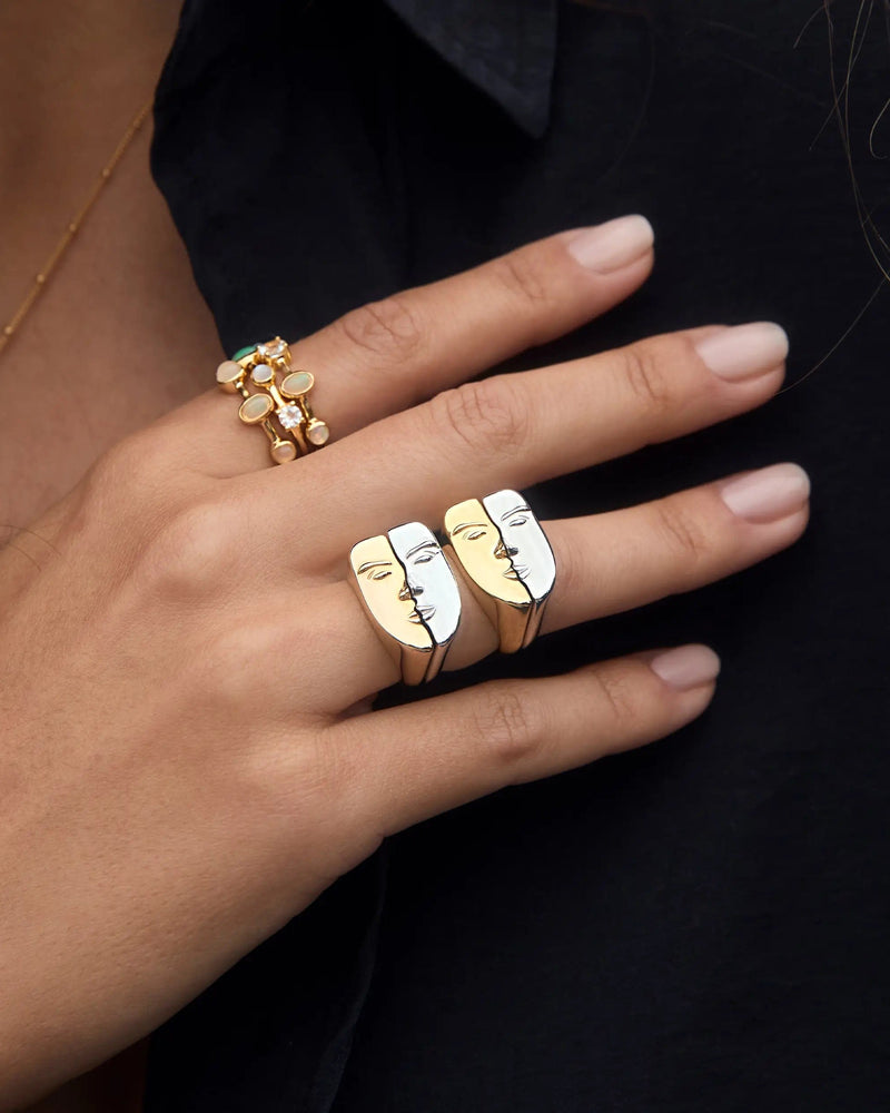 Close-up of a hand adorned with two Face to Face Rings designed by Awe Inspired, one in gold and black and the other in white and black. The person is dressed in a black garment and also wears a gold ring embellished with white stones.