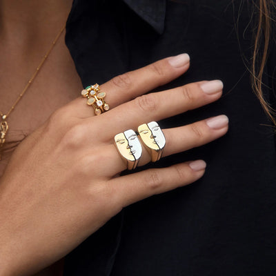 Close-up of a hand adorned with two Face to Face Rings designed by Awe Inspired, one in gold and black and the other in white and black. The person is dressed in a black garment and also wears a gold ring embellished with white stones.