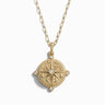 The Compass Locket Necklace by Awe Inspired is a gold necklace featuring a circular pendant adorned with intricate designs, centered with a pearl, and accented by four small diamonds, reminiscent of an exquisite compass rose.