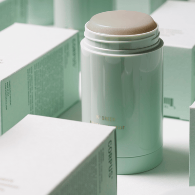 A green cylindrical deodorant container, known for its long-lasting formula and being gentle on underarms, is surrounded by multiple mint-colored product boxes. The product is the Third Rose Deodorant by Corpus.