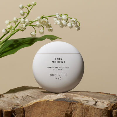 A round hand cream container with the text "This Moment Hand Care by Superegg" on a wooden surface, next to a branch with small white flowers. Infused with Murumuru and Shea Butters for ultimate hydration.